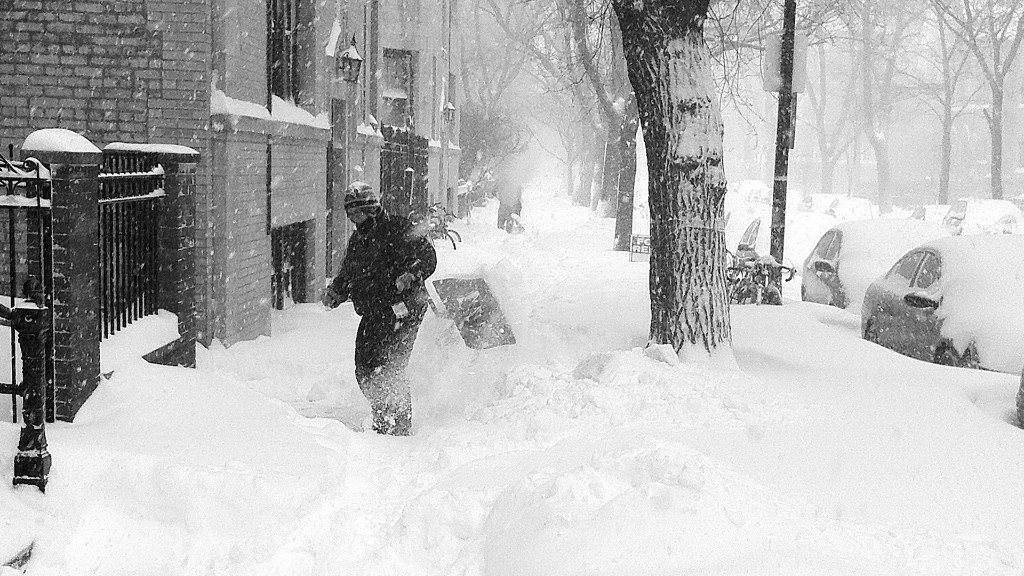 Sie sehen eine Person beim Schnee schaufeln in einer mit Häsuern gesämten Straße in einer Stadt.