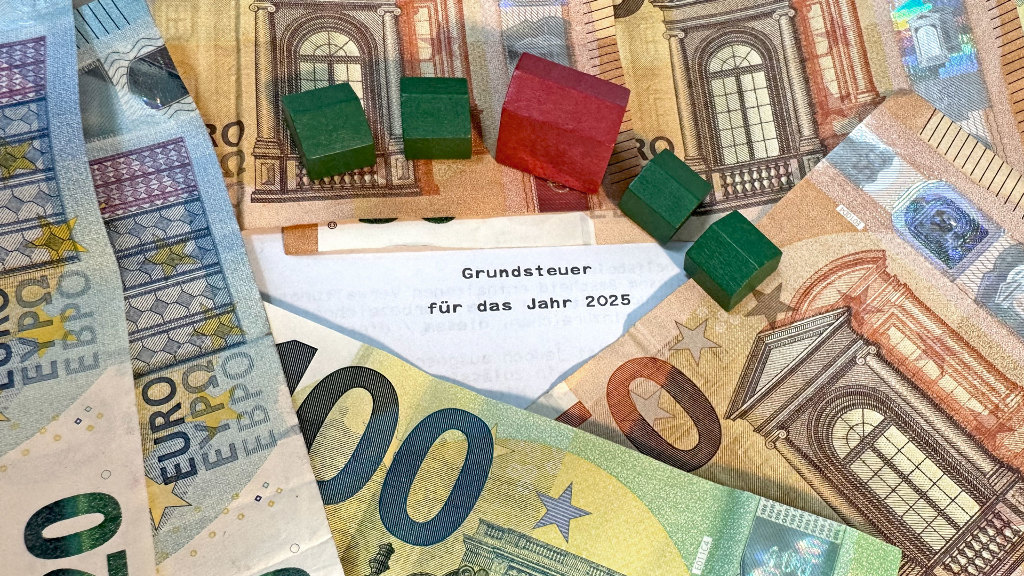 Sie sehen mehrere auf einem Grundsteuerbescheid verteilte Geldscheine, ergänzt um mehrere kleine Häuser.