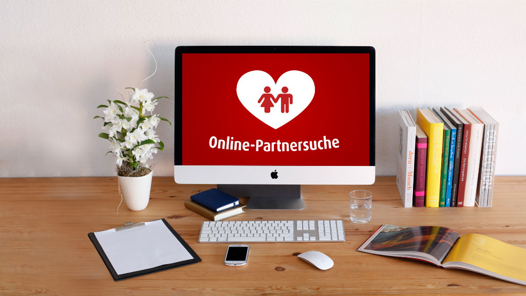 Sie sehen einen Computer auf einem Schreibtisch stehen, auf dessen Bildschirm ein Herz mit einem Pärchen darin zu sehen ist.