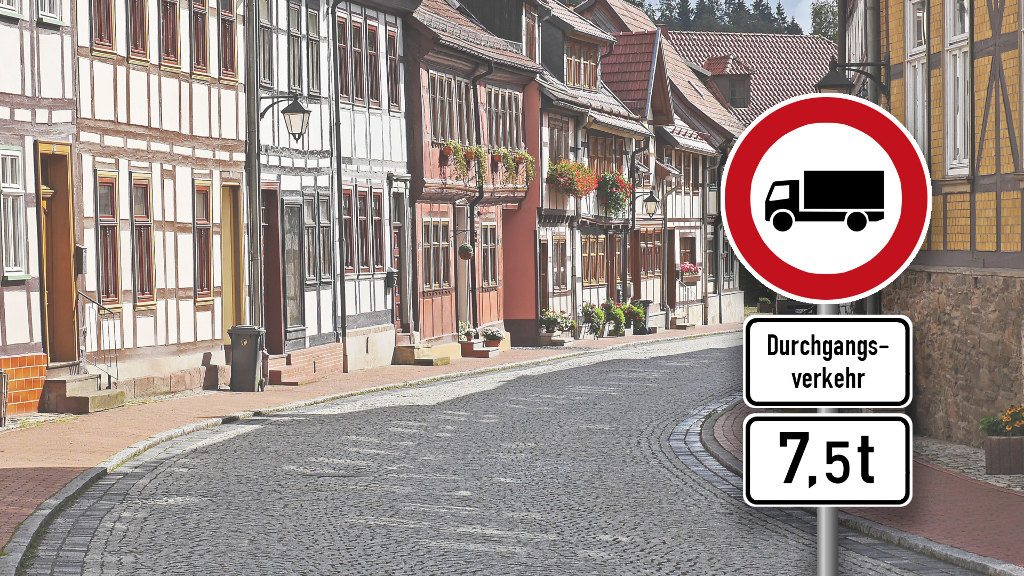 Sie sehen ein Verkehrsschild, welches die Durchfahrt für LKW über 7,5t Gesamtgewicht verbietet, vor der Kulisse einer Kleinstadt.