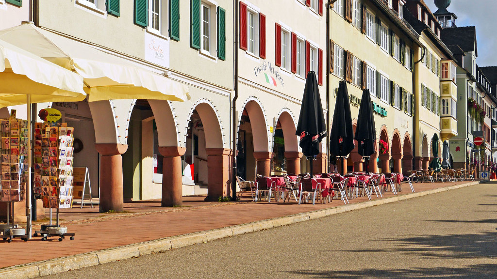 Sie sehen einen Fußweg in einer Stadt, auf dem Tische und Stühle eines Restaurants stehen.