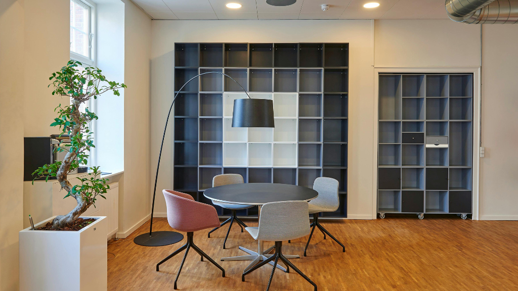 Sie sehen einen runden, schwarzen Holztisch mit 4 Stühlen vor einer Regalwand, wie es im Möbelhaus stehen könnte.