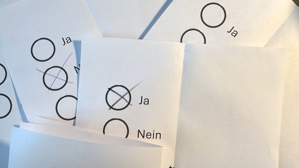 Sie sehen mehrere Stimmzettel mit einem Kreuz für &bdquo;ja&ldquo;, &bdquo;nein&ldquo; bzw. &bdquo;Enthaltung&ldquo; und einen teilweise zugeklappten Stimmzettel.