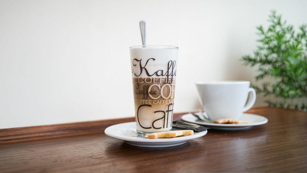 Sie sehen ein Glas Latte Macchiato und eine Kaffeetasse auf einer kleinen Theke stehen.