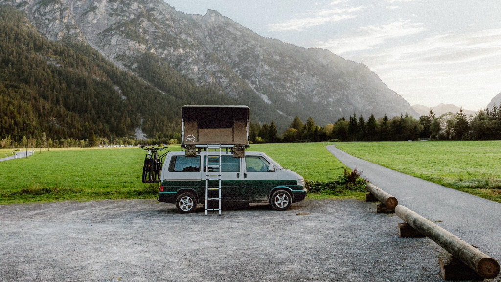 Sie sehen einen VW Bus mit Dachzelt, geparkt an einer Landstraße mit Bergen im Hintergrund.