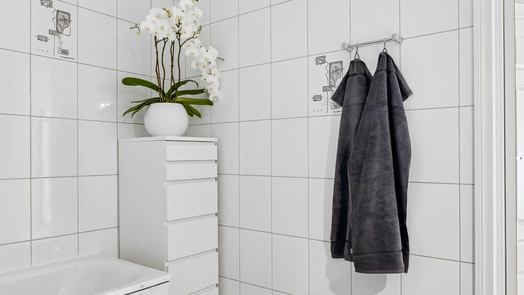 Sie sehen ein modernes, minimalistisches Badezimmer mit gefliesten Wänden, einem Schrank und darauf eine weiße Orchidee in einer Vase.