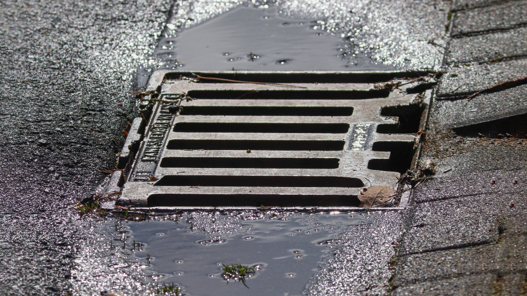 Sie sehen einen Gulli in einer teilweise gepflasterten Straße, an dessen Rand sich Regenwasser sammelt.