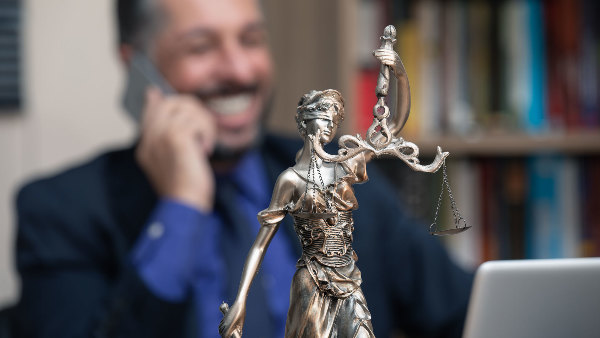 Sie sehen ein Justizia-Figur und im Hintergrund einen Mann am Telefon.