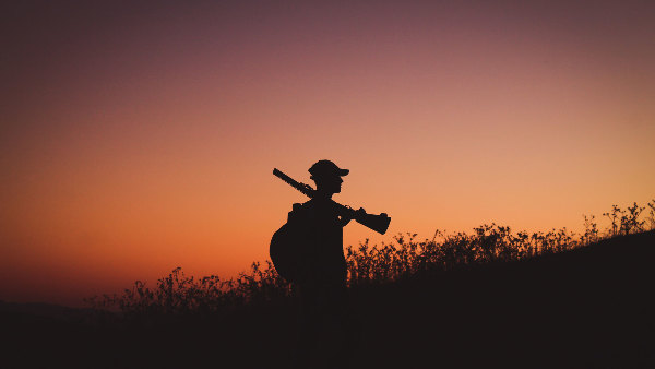 Sie sehen einen Jäger, der in der Dämmerung mit geschultertem Gewehr einen Hügel hinaufgeht.