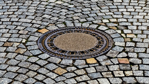 Sie sehen ein Kopfsteinpflaster mit einem eingelassenen Gullideckel.