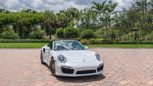 Sie sehen einen weißen Porsche Turbo S Cabriolet auf einem, von Bäumen umrahmten, Platz stehen.
