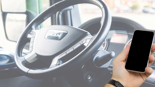 Sie sehen das Lenkrad eines LKW oder Busses und eine Handy, welche ein Smartphone hält.