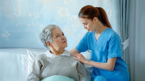 Sie sehen eine Pflegekraft, die mit einer älteren Dame spricht.