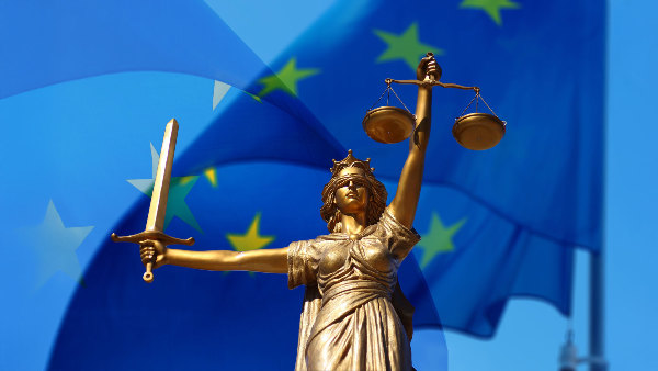 Sie sehen eine Statue der Justizia vor einer Europa-Fahne.
