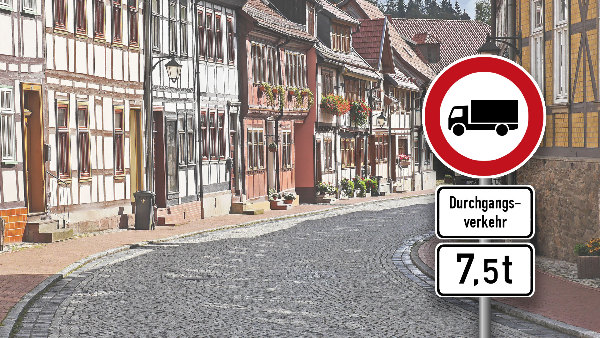 Sie sehen ein Verkehrsschild, welches die Durchfahrt für LKW über 7,5t Gesamtgewicht verbietet, vor der Kulisse einer Kleinstadt.
