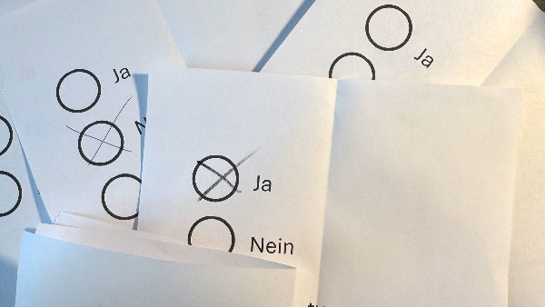 Sie sehen mehrere Stimmzettel mit einem Kreuz für &bdquo;ja&ldquo;, &bdquo;nein&ldquo; bzw. &bdquo;Enthaltung&ldquo; und einen teilweise zugeklappten Stimmzettel.