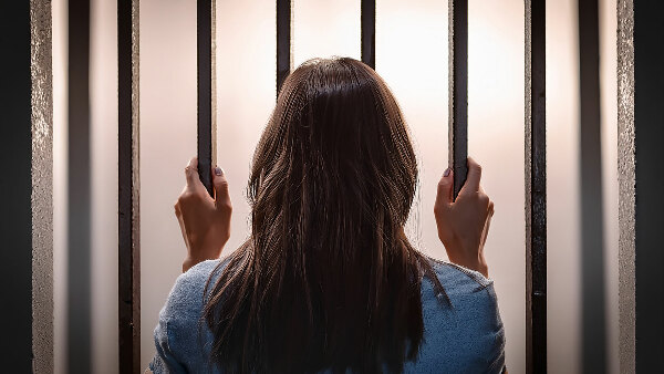 Sie sehen eine junge Frau von hinten, die Gitterstäbe in einem Gefängnis umfasst.