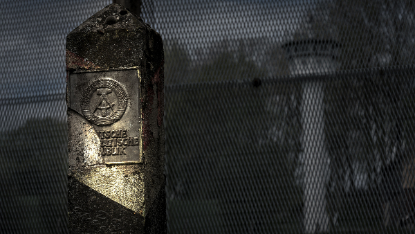 Sie sehen einen Grenzzaun mit einem Wachturm im Hintergrund und einem Pfahl mit Symbolik der Deutschen Demokratischen Republik.