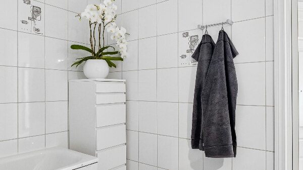 Sie sehen ein modernes, minimalistisches Badezimmer mit gefliesten Wänden, einem Schrank und darauf eine weiße Orchidee in einer Vase.