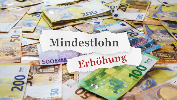 Sie sehen viele verteilte Euro-Banknoten, ergänzt um zwei Papierschnipsel mit den Worten &bdquo;Mindestlohn&ldquo; und &bdquo;Erhöhung&ldquo; darauf.