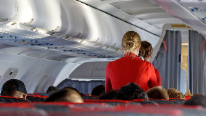 Sie sehen den besetzten Innenraum eines Linienflugzeugs und zwei Stewardessen im Gang zwischen den Sitzen.