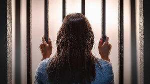Sie sehen eine junge Frau von hinten, die Gitterstäbe in einem Gefängnis umfasst.