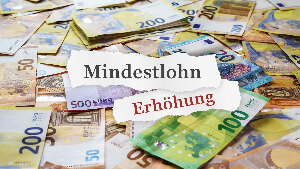 Sie sehen viele verteilte Euro-Banknoten, ergänzt um zwei Papierschnipsel mit den Worten &bdquo;Mindestlohn&ldquo; und &bdquo;Erhöhung&ldquo; darauf.
