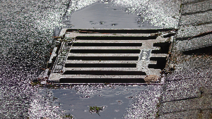 Sie sehen einen Gulli in einer teilweise gepflasterten Straße, an dessen Rand sich Regenwasser sammelt.